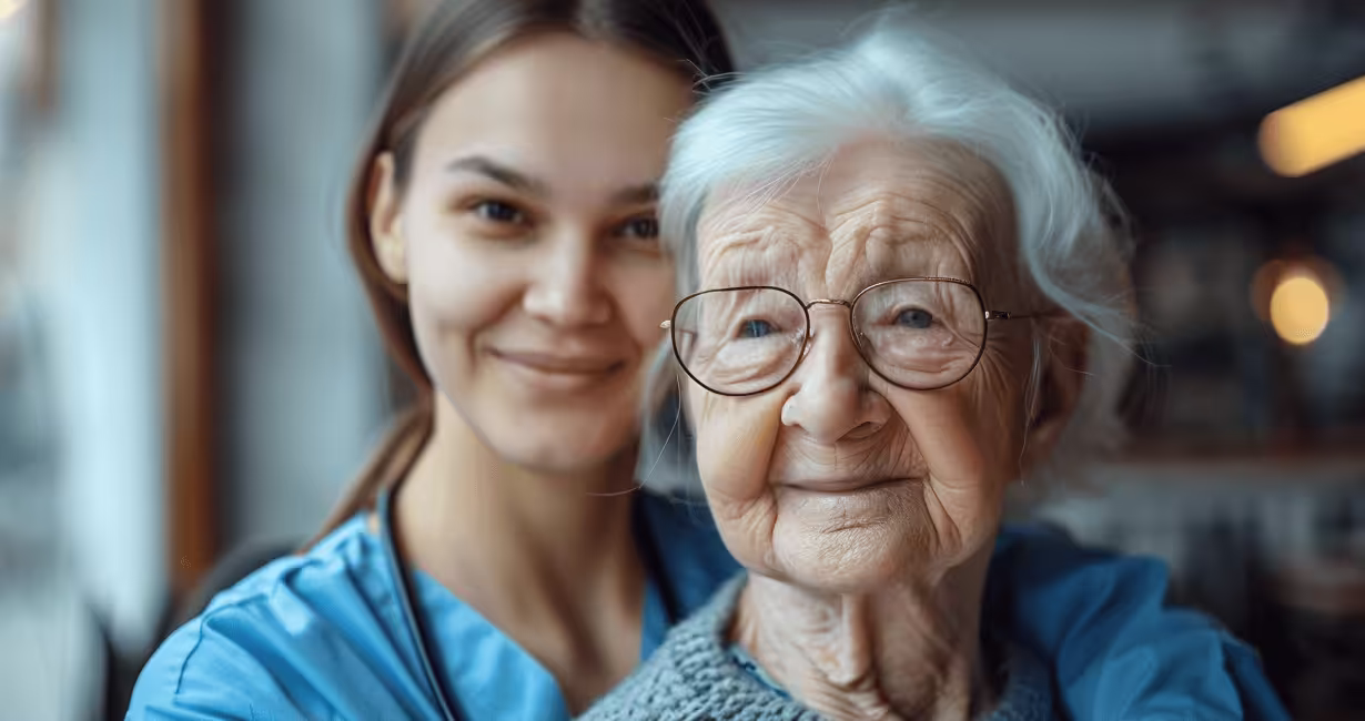 Eine Pflegekraft umarmt eine ältere Frau und lächelt in die Kamera.